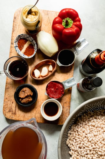 Overhead flat-lay of all the baked bean ingredients prepped on a cutting board and counter: tomato paste, maple syrup, vegan Worcestershire sauce, miso, smoked paprika, onion powder, a yellow onion, a red bell pepper, garlic cloves in a small bowl, vegetable stock, liquid smoke, balsamic vinegar, Dijon mustard, and a colander of soaked white beans.