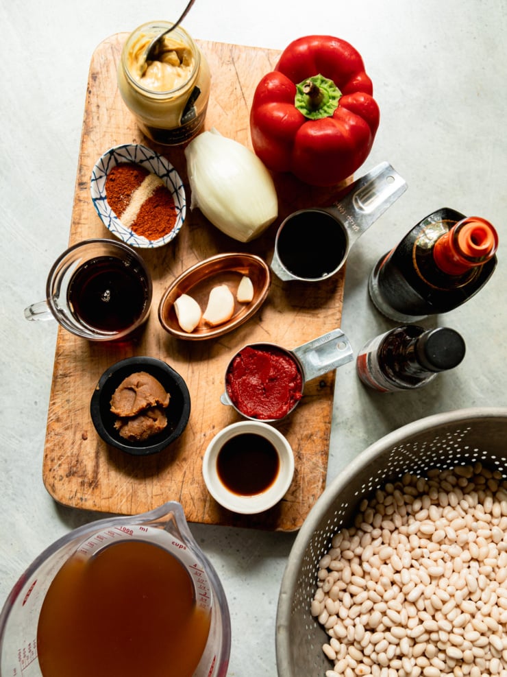 Overhead flat-lay of all the baked bean ingredients prepped on a cutting board and counter: tomato paste, maple syrup, vegan Worcestershire sauce, miso, smoked paprika, onion powder, a yellow onion, a red bell pepper, garlic cloves in a small bowl, vegetable stock, liquid smoke, balsamic vinegar, Dijon mustard, and a colander of soaked white beans.