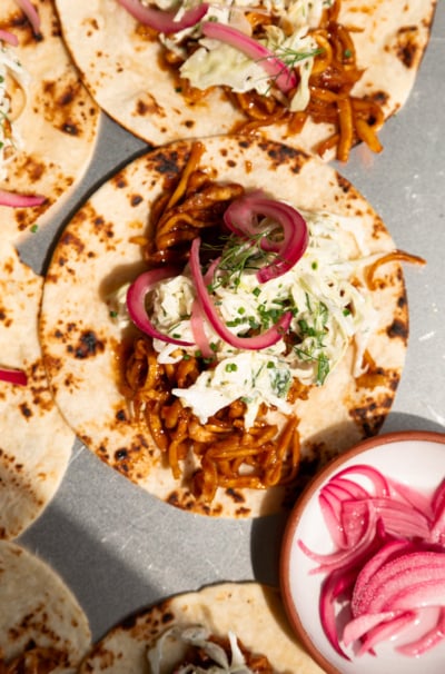 Overhead shot of assembled tacos on charred flour tortillas, topped with saucy shredded BBQ tofu, creamy cabbage slaw, pickled red onions, and fresh dill and chives.