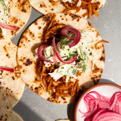 Overhead shot of assembled tacos on charred flour tortillas, topped with saucy shredded BBQ tofu, creamy cabbage slaw, pickled red onions, and fresh dill and chives.