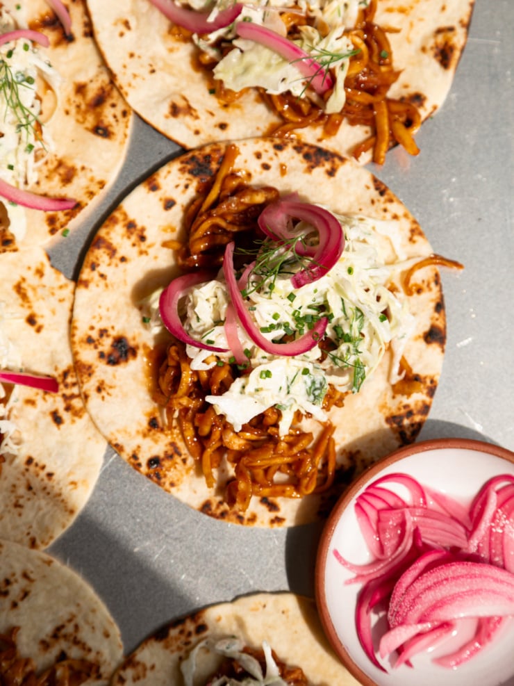 Overhead shot of assembled tacos on charred flour tortillas, topped with saucy shredded BBQ tofu, creamy cabbage slaw, pickled red onions, and fresh dill and chives.