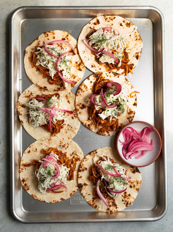 Six charred flour tortillas topped with BBQ tofu and yogurt ranch slaw arranged on a metal baking sheet, with a small bowl of pickled red onions on the side.