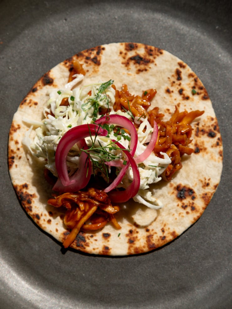 A single taco on a dark plate, filled with shredded BBQ tofu, cabbage slaw, pickled red onions, dill, and chives, viewed from above.
