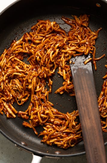 Shredded smoked tofu coated in BBQ sauce sizzling in a nonstick skillet with a wooden spatula.
