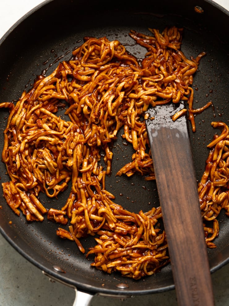 Shredded smoked tofu coated in BBQ sauce sizzling in a nonstick skillet with a wooden spatula.