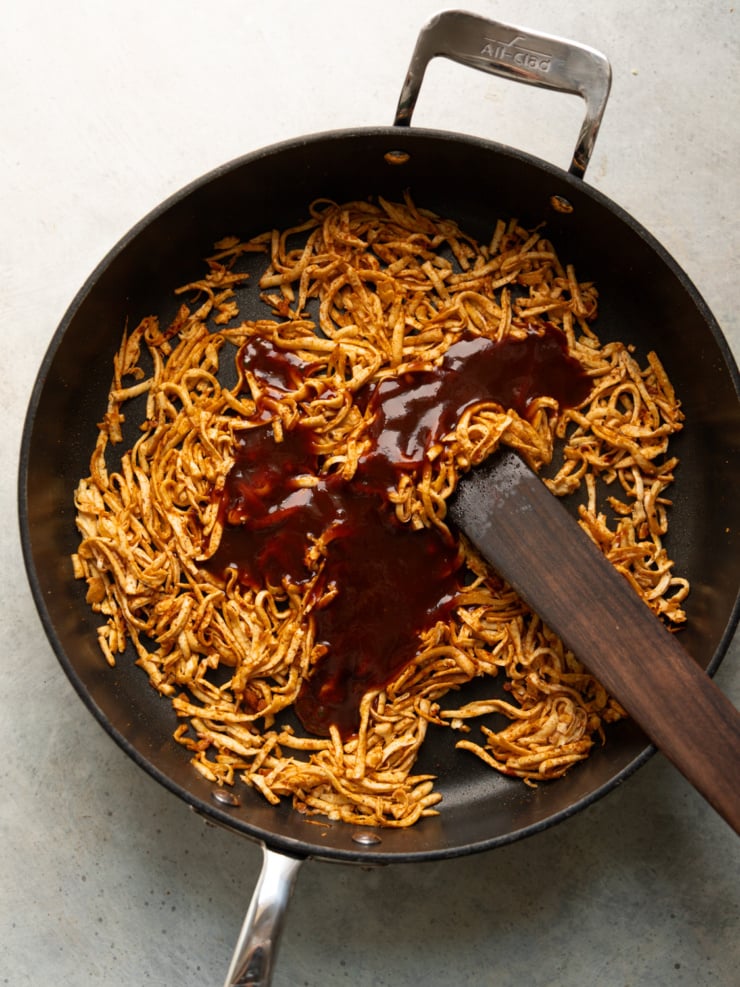 Seasoned shredded tofu in a skillet with BBQ sauce being poured over the top before stirring together.