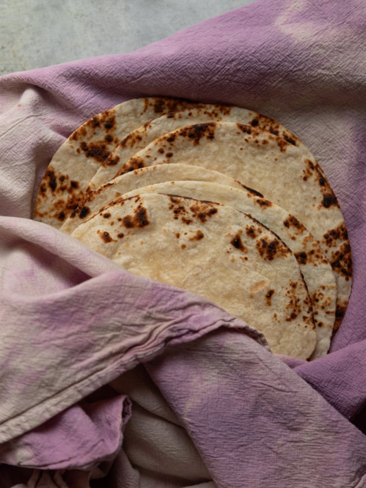 A stack of charred flour tortillas resting on a lavender-dyed linen cloth.