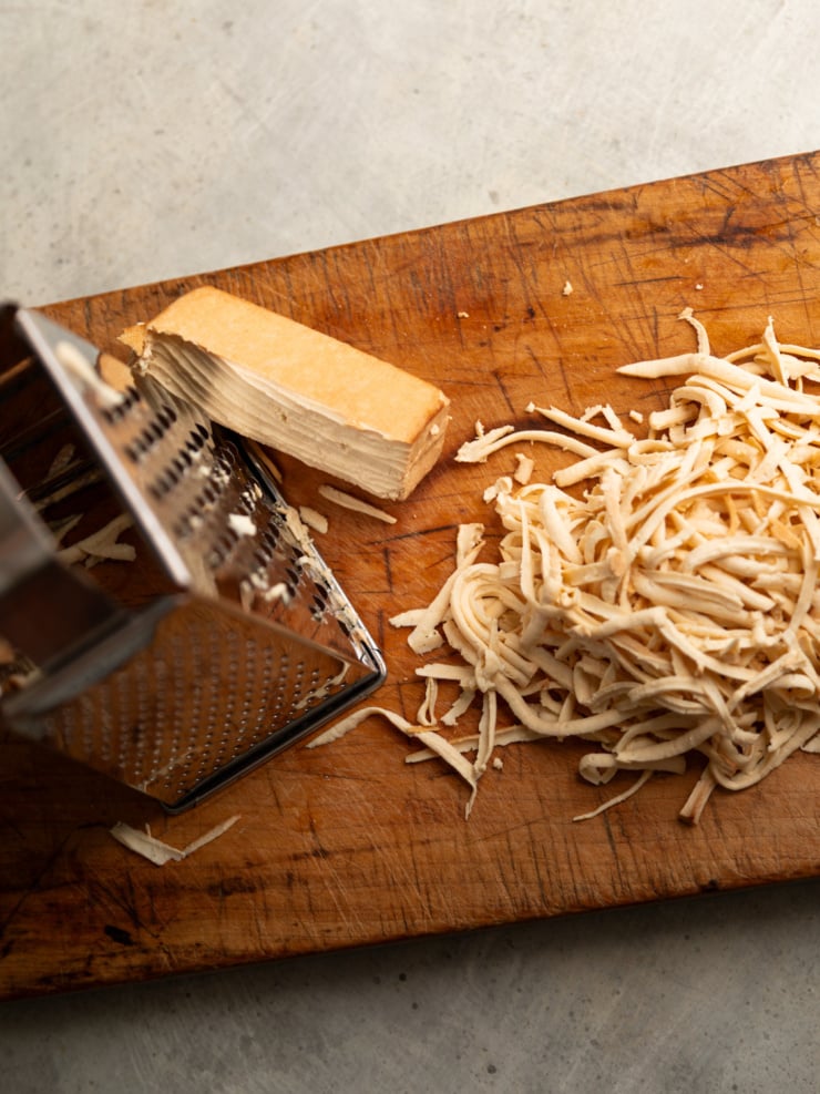 A block of tofu being grated into thin shreds on a wooden cutting board, with a pile of shredded tofu beside the grater.