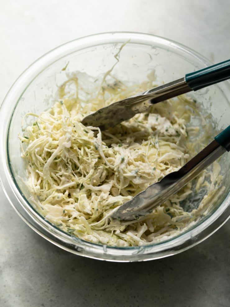 A glass bowl of shredded cabbage being tossed with creamy yogurt ranch dressing using metal tongs.