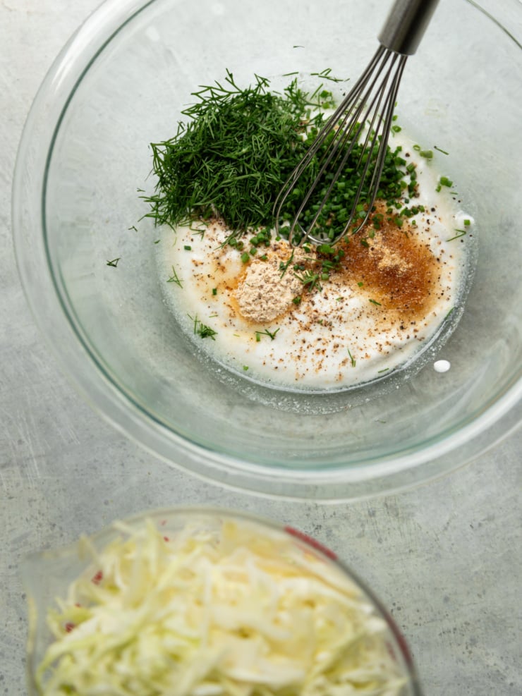 A glass bowl of yogurt ranch dressing being whisked together with fresh dill, chives, and spices, with a bowl of shredded cabbage in the foreground.