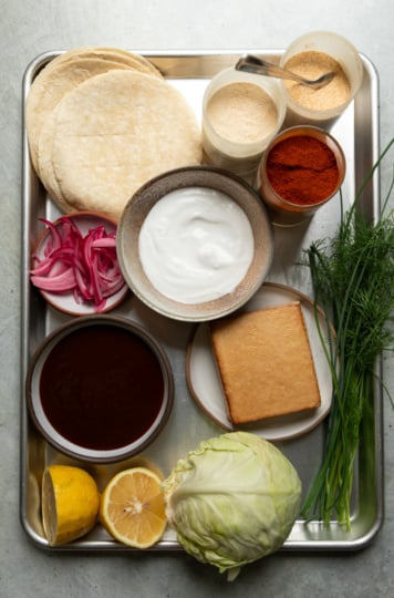Overhead shot of all the taco ingredients laid out on a metal tray: flour tortillas, spices, vegan yogurt, BBQ sauce, a block of smoked tofu, pickled red onions, fresh dill and chives, lemon halves, and a head of cabbage.