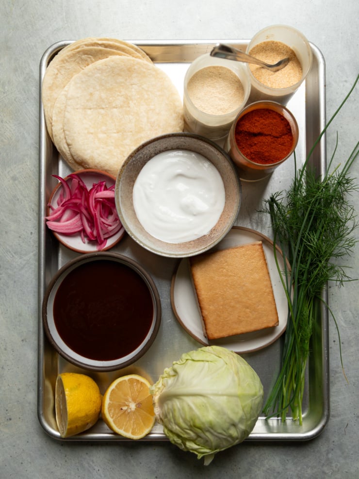 Overhead shot of all the taco ingredients laid out on a metal tray: flour tortillas, spices, vegan yogurt, BBQ sauce, a block of smoked tofu, pickled red onions, fresh dill and chives, lemon halves, and a head of cabbage.
