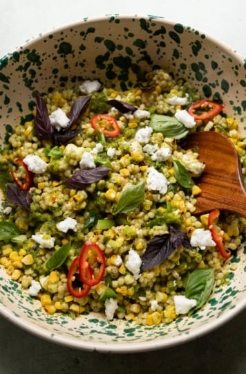 Overhead shot of charred corn couscous salad in a cream and green speckled bowl, topped with vegan feta crumbles, sliced Fresno chilies, purple and green basil leaves, with a wooden spoon resting in the bowl.