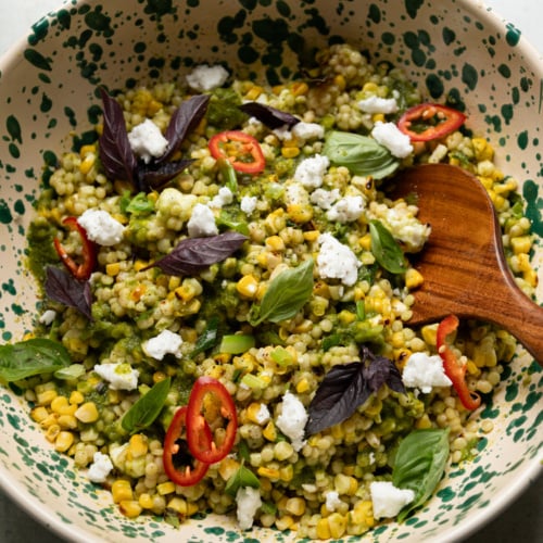 Overhead shot of charred corn couscous salad in a cream and green speckled bowl, topped with vegan feta crumbles, sliced Fresno chilies, purple and green basil leaves, with a wooden spoon resting in the bowl.