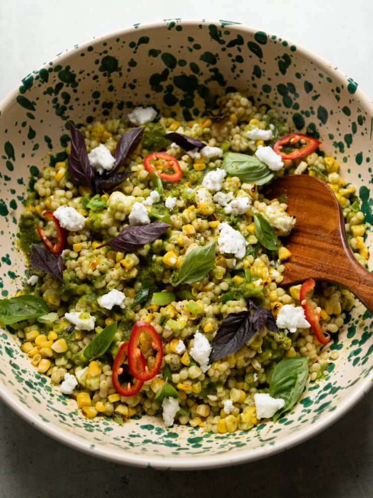 Overhead shot of charred corn couscous salad in a cream and green speckled bowl, topped with vegan feta crumbles, sliced Fresno chilies, purple and green basil leaves, with a wooden spoon resting in the bowl.