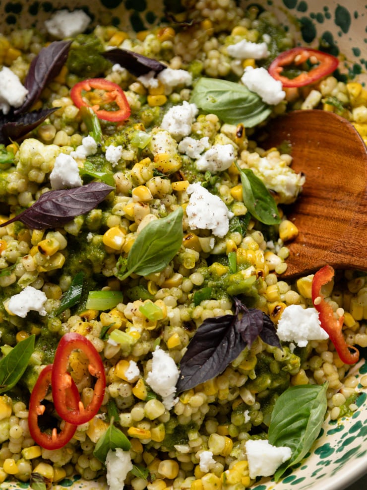 Close-up of charred corn couscous salad showing pearl couscous, charred corn kernels, green onion, vegan feta, purple basil, and sliced Fresno chili rings.