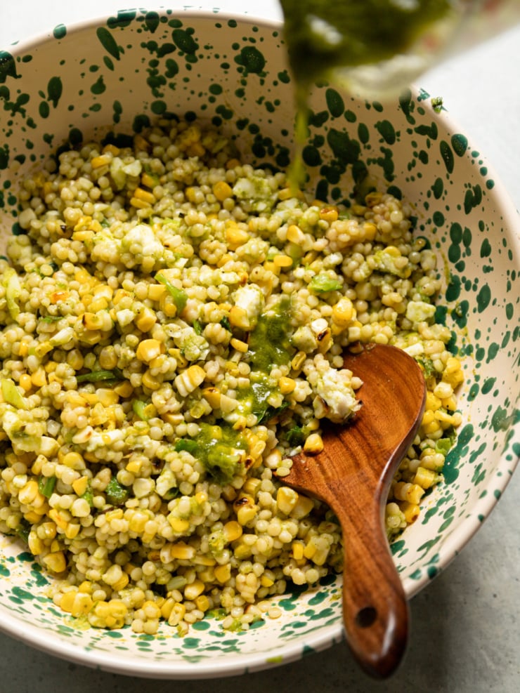 Basil lime dressing being poured over charred corn couscous salad in a speckled ceramic bowl with a wooden spoon.