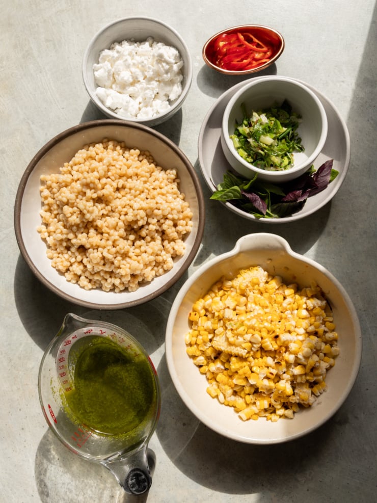 Overhead shot of charred corn couscous salad ingredients prepped in bowls, including cooked pearl couscous, charred corn, vegan feta, sliced Fresno chili, chopped green onion, basil leaves, and basil lime dressing.