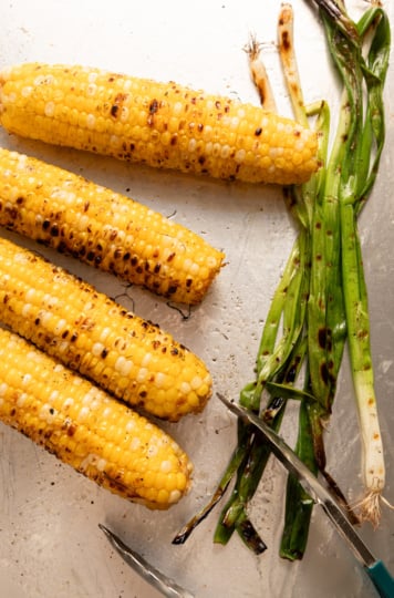 Charred corn on the cob and grilled green onions resting on a baking sheet with metal tongs.