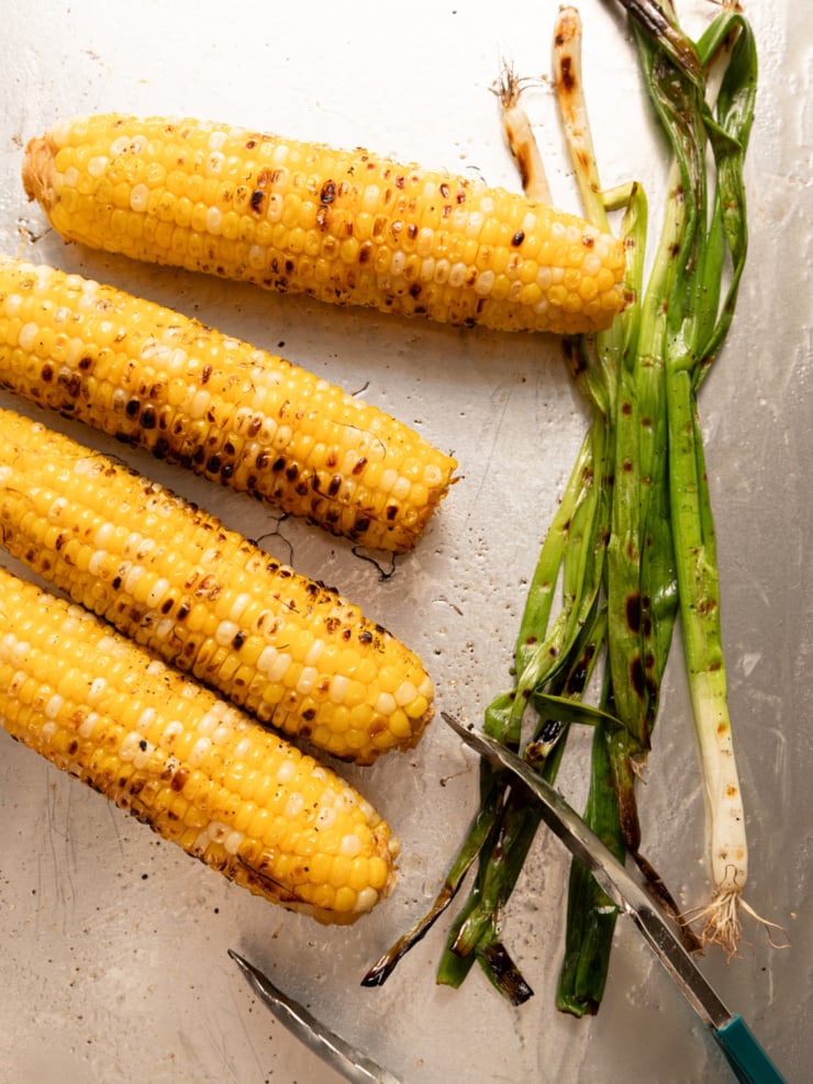 Charred corn on the cob and grilled green onions resting on a baking sheet with metal tongs.