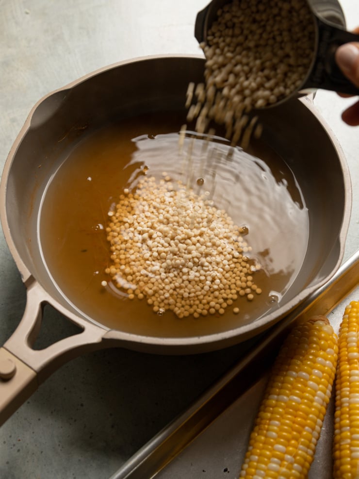 Pearl couscous being poured into a deep skillet of broth for cooking.