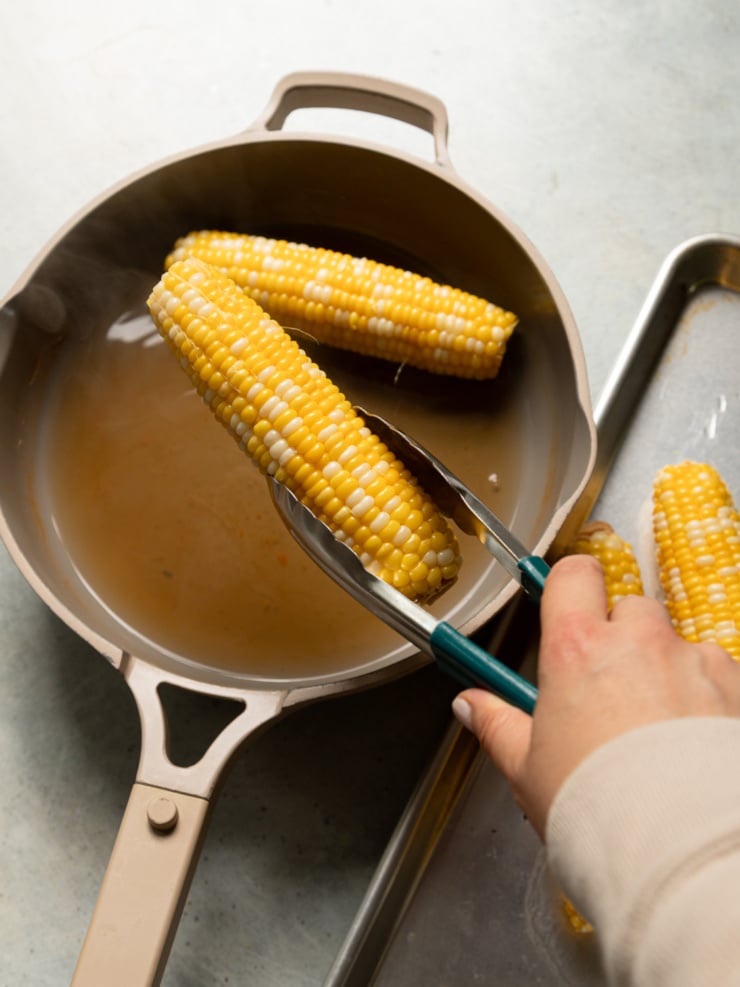Tongs lifting a charred ear of corn out of a pot of hot vegetable stock.