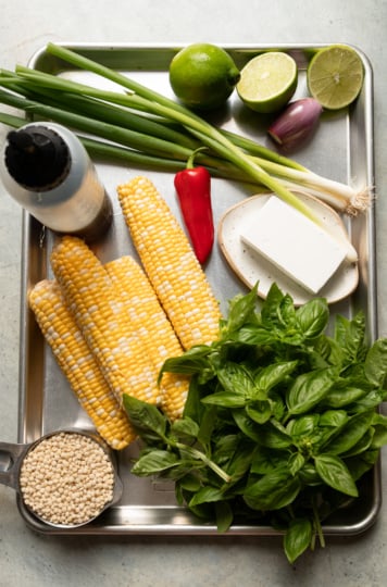 Overhead shot of ingredients for charred corn couscous salad arranged on a metal tray, including corn on the cob, green onions, a Fresno chili, limes, shallot, vegan feta, fresh basil, pearl couscous, and a squeeze bottle of agave nectar.