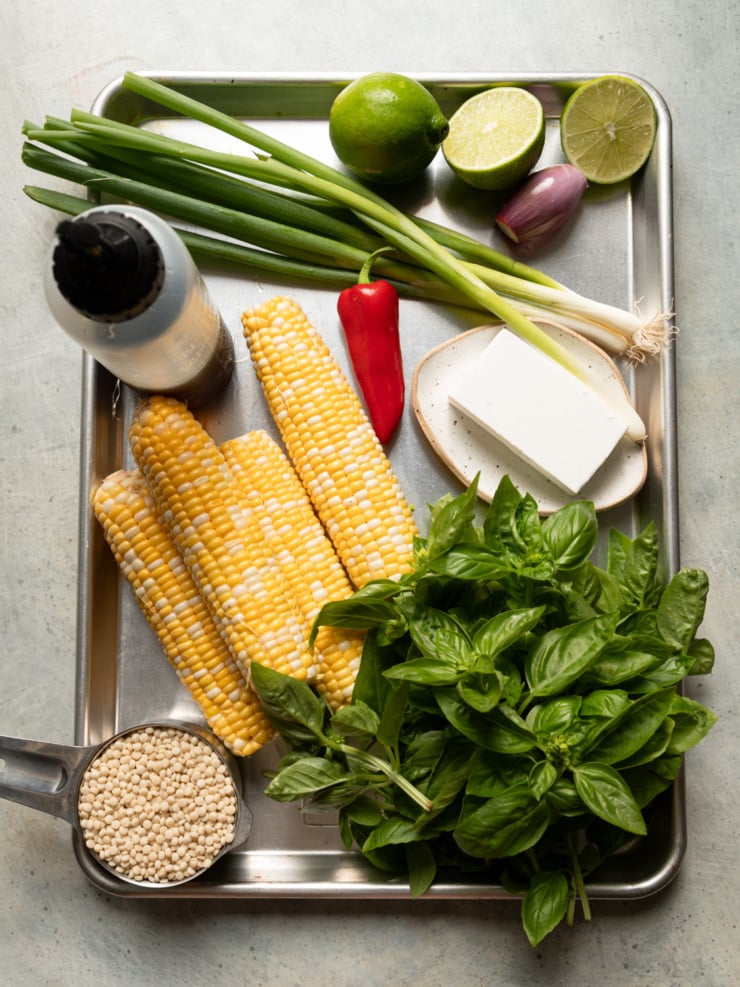 Overhead shot of ingredients for charred corn couscous salad arranged on a metal tray, including corn on the cob, green onions, a Fresno chili, limes, shallot, vegan feta, fresh basil, pearl couscous, and a squeeze bottle of agave nectar.