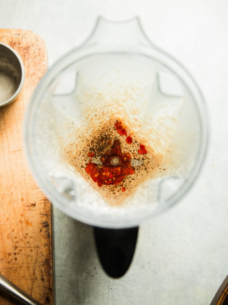 Empty blender jar with minced Calabrian chilis, cracked black pepper, and garlic at the bottom, ready to blend into dip.