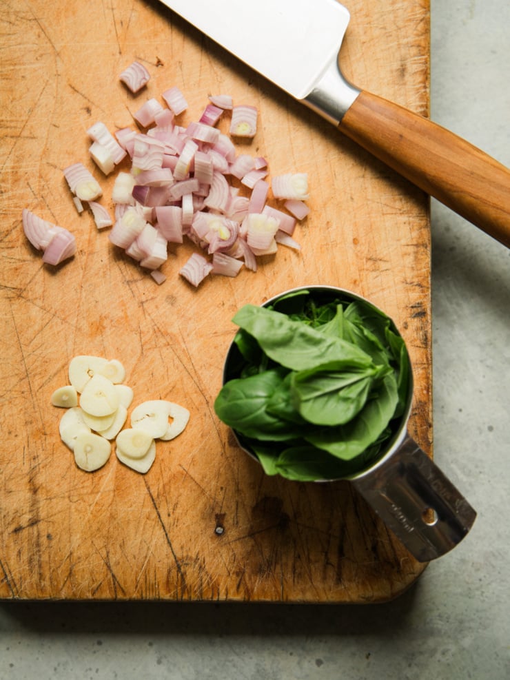 Diced shallot, sliced garlic, and a cup of fresh basil on a wooden cutting board, prepped for garlic white bean dip.