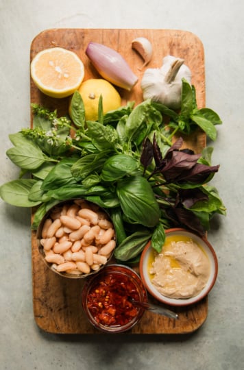 Overhead view of ingredients for garlic white bean dip: white beans, fresh basil, garlic, shallot, lemon, raw smooth cashew butter, and Calabrian chili paste.