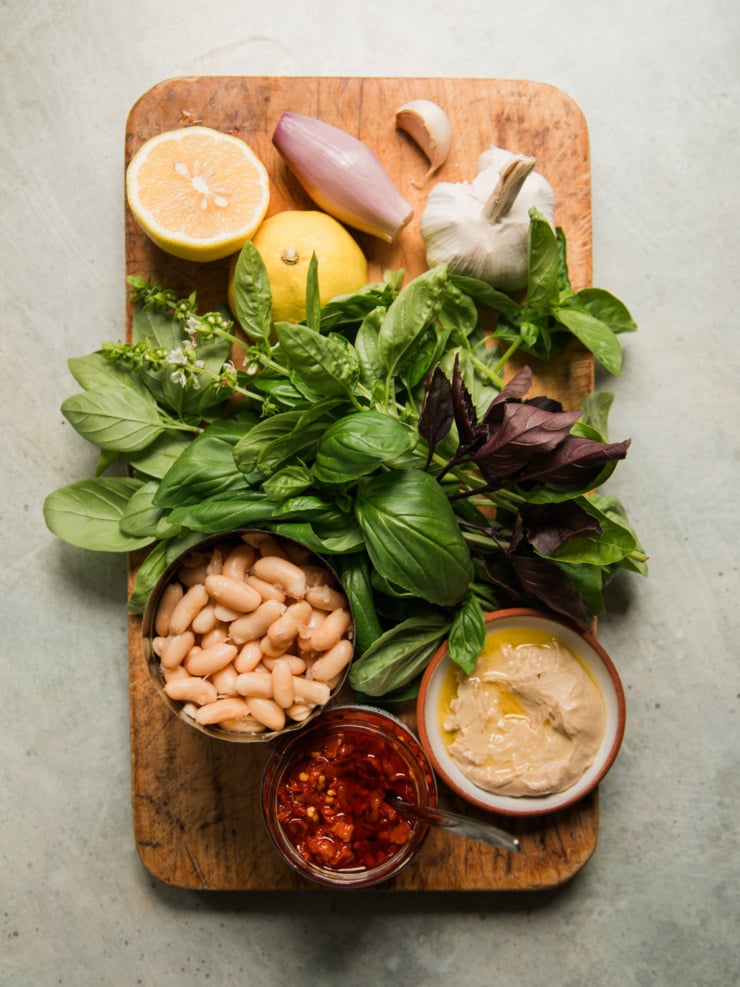 Overhead view of ingredients for garlic white bean dip: white beans, fresh basil, garlic, shallot, lemon, raw smooth cashew butter, and Calabrian chili paste.