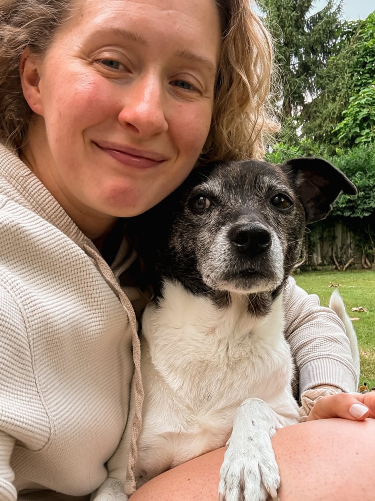 Laura smiling cheek-to-cheek with an Cleo, older black and white dog, sitting together outside in a backyard with grass and trees in the background.