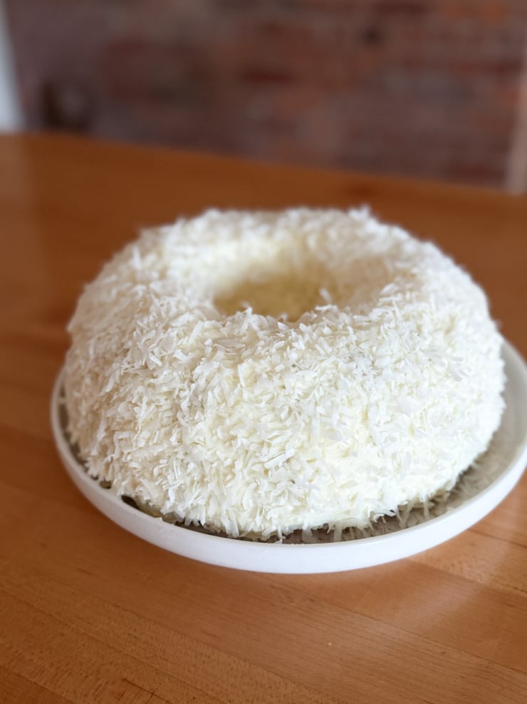 A round coconut-covered cake on a white plate, fully coated in shredded coconut, sitting on a wooden table with a blurred brick wall behind it.