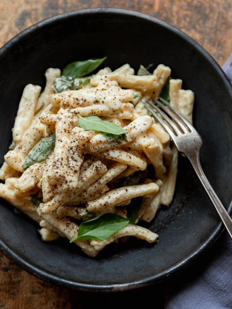 Overhead shot of casarecce pasta in a creamy caramelized onion and yellow squash sauce in a dark ceramic bowl, garnished with fresh basil leaves and a fork resting on the side.