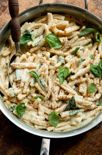 Overhead view of creamy caramelized onion and yellow squash pasta in a stainless steel skillet, topped with fresh basil leaves and a dusting of black pepper, with a serving spoon.