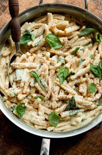Overhead view of creamy caramelized onion and yellow squash pasta in a stainless steel skillet, topped with fresh basil leaves and a dusting of black pepper, with a serving spoon.