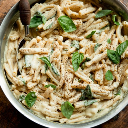 Overhead view of creamy caramelized onion and yellow squash pasta in a stainless steel skillet, topped with fresh basil leaves and a dusting of black pepper, with a serving spoon.