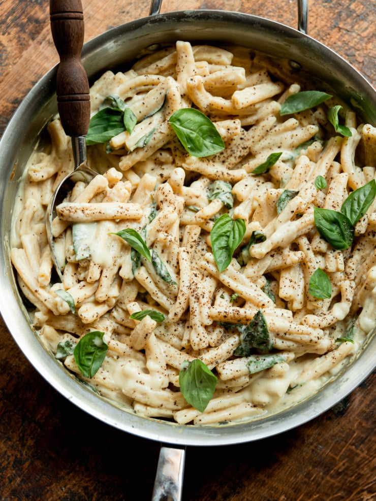 Overhead view of creamy caramelized onion and yellow squash pasta in a stainless steel skillet, topped with fresh basil leaves and a dusting of black pepper, with a serving spoon.