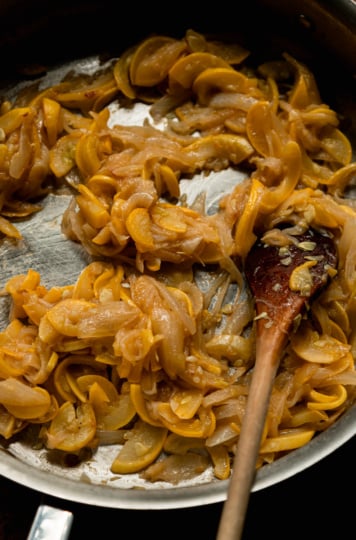Caramelized onions and sliced yellow squash cooking together in a stainless steel skillet, being stirred with a wooden spoon.