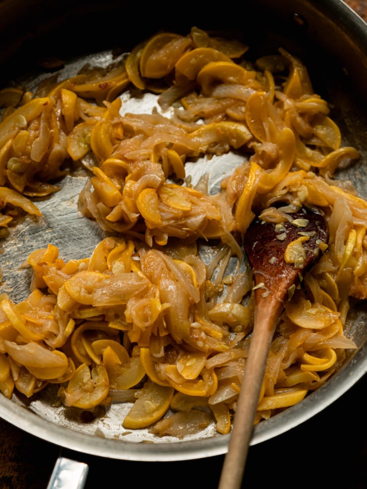 Caramelized onions and sliced yellow squash cooking together in a stainless steel skillet, being stirred with a wooden spoon.