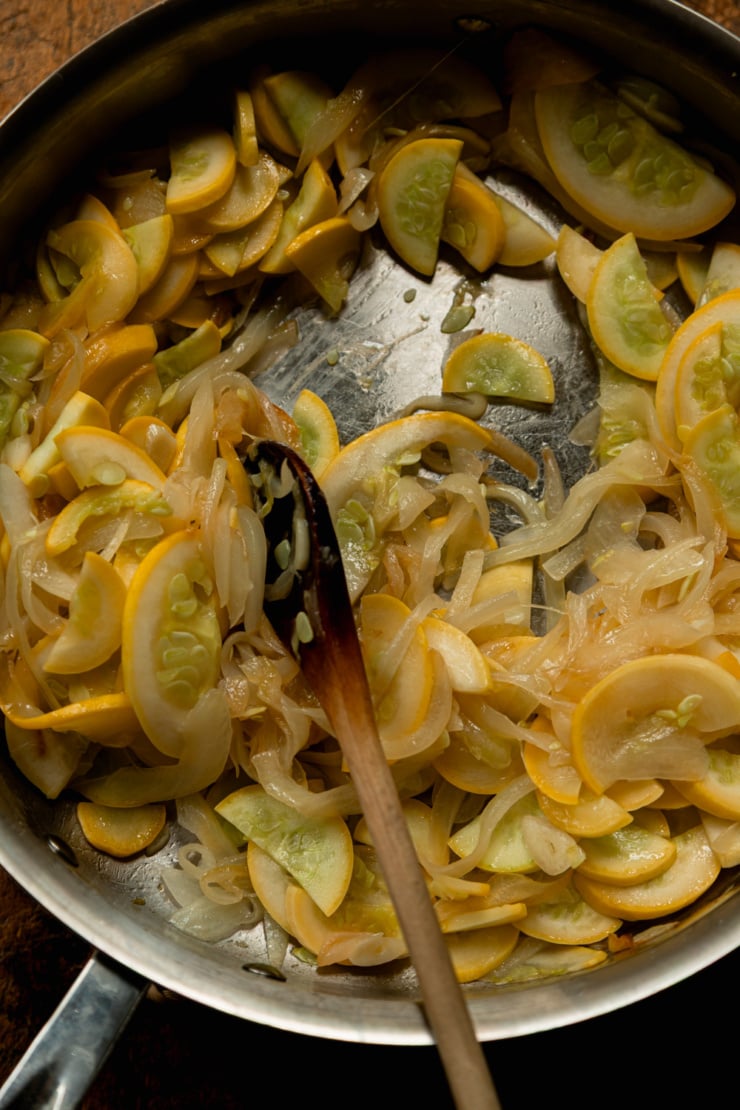 Overhead view of sliced yellow squash added to caramelized onions in a stainless steel skillet, with a wooden spoon.