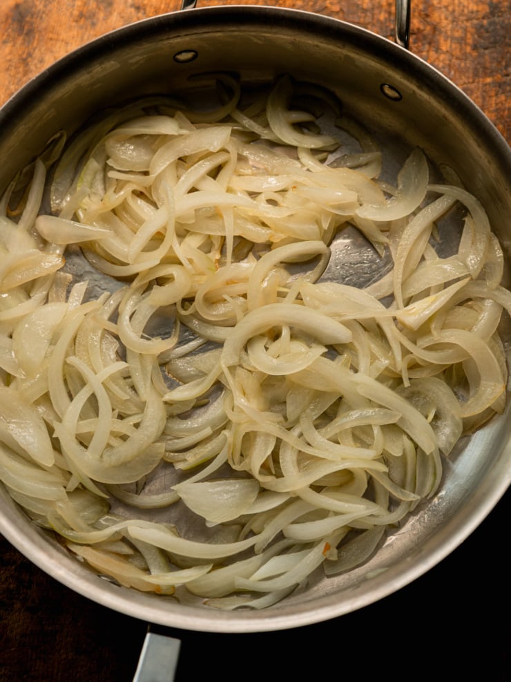 Overhead view of softened, translucent sliced onions cooking in a stainless steel skillet.