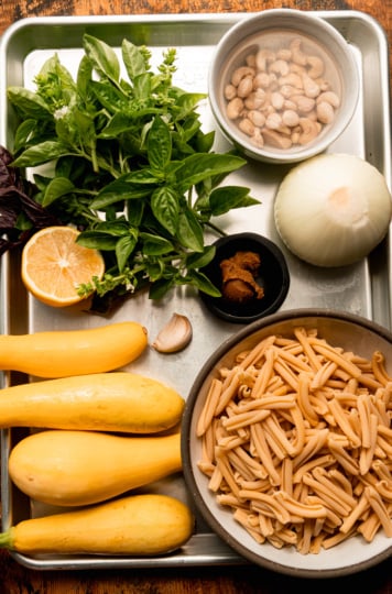 Overhead shot of ingredients laid out on a sheet pan for caramelized onion and squash pasta: fresh basil, a halved lemon, a bowl of soaking cashews, a whole onion, a small bowl of miso paste, a garlic clove, four yellow squash, and a bowl of dried casarecce pasta.
