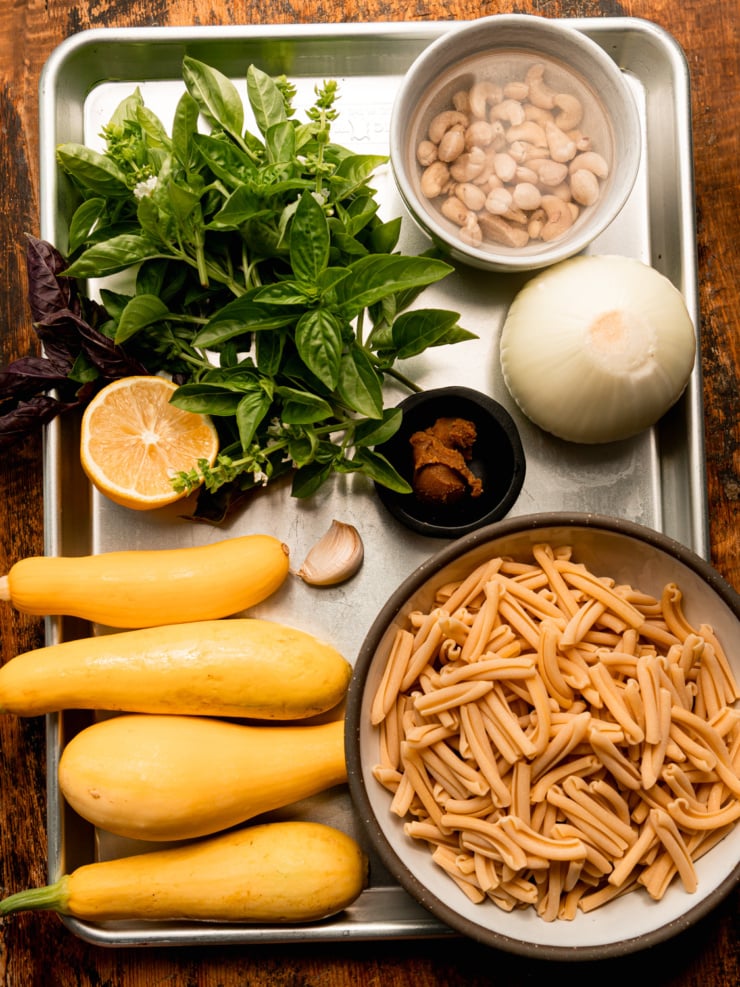 Overhead shot of ingredients laid out on a sheet pan for caramelized onion and squash pasta: fresh basil, a halved lemon, a bowl of soaking cashews, a whole onion, a small bowl of miso paste, a garlic clove, four yellow squash, and a bowl of dried casarecce pasta.