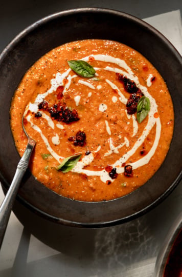 Overhead shot of creamy red lentil tomato soup in a dark, rustic ceramic bowl. The vibrant orange-red soup is swirled with white tahini drizzle and topped with dollops of dark, glistening chili crisp, fresh basil leaves, and a scattering of red pepper flakes and cracked black pepper. A silver spoon rests in the bowl on the left side, and the edge of a second bowl of soup is visible in the bottom right corner.