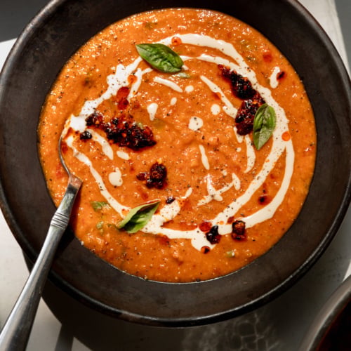 Overhead shot of creamy red lentil tomato soup in a dark, rustic ceramic bowl. The vibrant orange-red soup is swirled with white tahini drizzle and topped with dollops of dark, glistening chili crisp, fresh basil leaves, and a scattering of red pepper flakes and cracked black pepper. A silver spoon rests in the bowl on the left side, and the edge of a second bowl of soup is visible in the bottom right corner.