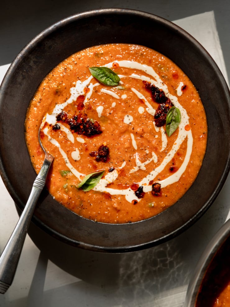 Overhead shot of creamy red lentil tomato soup in a dark, rustic ceramic bowl. The vibrant orange-red soup is swirled with white tahini drizzle and topped with dollops of dark, glistening chili crisp, fresh basil leaves, and a scattering of red pepper flakes and cracked black pepper. A silver spoon rests in the bowl on the left side, and the edge of a second bowl of soup is visible in the bottom right corner.