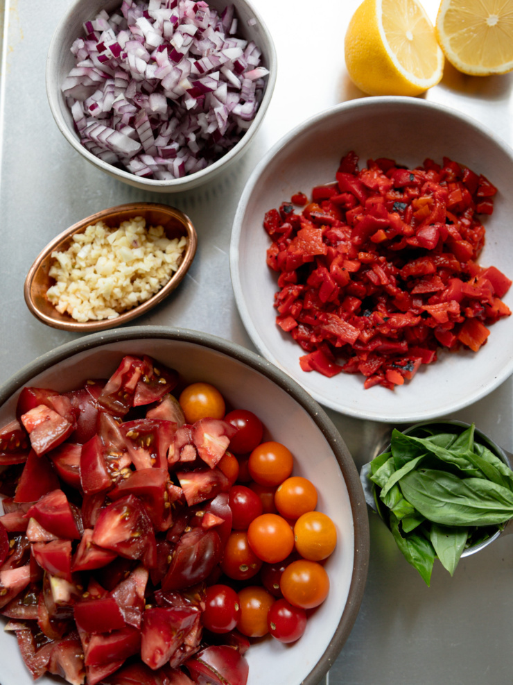 Overhead shot of mise en place for red lentil tomato soup, arranged in small bowls on a metal tray: diced red onion, minced garlic, chopped charred roasted red peppers, and a mix of chopped heirloom tomatoes with whole cherry and grape tomatoes, plus a small bowl of fresh basil leaves and two lemon halves in the corner.
