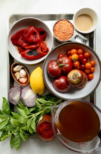 Overhead flat-lay of all ingredients for red lentil tomato soup arranged on a metal baking tray: whole roasted red peppers, a measuring cup of dried red lentils, a small cup of tahini, whole heirloom and cherry tomatoes in a bowl, garlic cloves, a whole lemon, halved red onion, a bunch of fresh basil, a small dish of smoked paprika or chili powder, and a large measuring cup of vegetable broth.